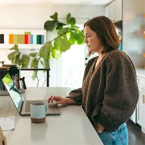Mujer emprendedora usando su portátil en casa, rodeada de un ambiente de cocina y trabajo.